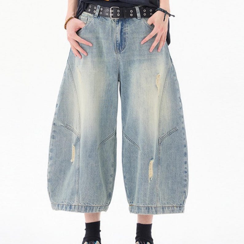 May include: Light blue denim culottes with a distressed, faded wash. The wide-leg pants feature a high waist, a black belt with silver-tone grommets, and ripped details. The culottes are a relaxed fit and have a unique, voluminous shape.