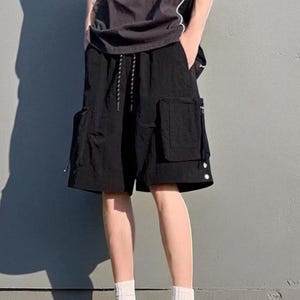 Multi Pocket Cargo Shorts | Men Baggy Streetwear Shorts | Casual Summer Outfit | Oversized Hip Hop Shorts | Gift for Him
.
Person wearing loose-fit black cargo shorts with multiple flap and zipper pockets, standing outdoors on a grassy field under a clear sky.