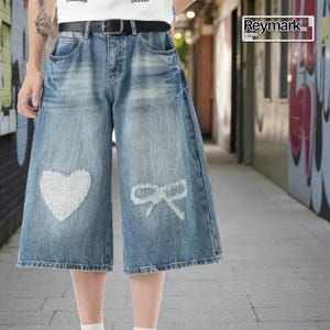 May include: Blue denim culottes with a distressed heart and bow design. The wide-leg pants have a faded wash and a black belt. The brand name "Reymark" is visible in the upper right corner.