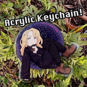 May include: A circular acrylic keychain featuring an illustration of a blonde character in a black coat and brown boots holding a coffee cup against a dark purple starry background. The text Acrylic Keychain! is printed in white above the object.