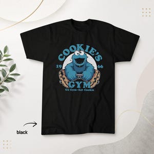 May include: A black short-sleeve t-shirt featuring a graphic of a blue monster driving with chocolate chip cookies as wheels. The text reads COOKIE'S GYM, 1966, and Go Gym Eat Cookie in light blue. The design is centred on the chest area.
