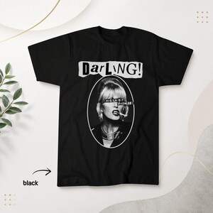 May include: A black short-sleeved t-shirt featuring a white graphic print of a person with a cigarette. Above the oval portrait, the text reads "DarLING!" and a black bar across the eyes contains the white text "ABSOLUTELY FABULOUS".