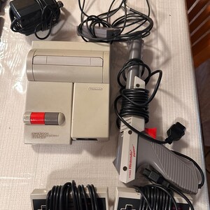 May include: A vintage Nintendo Entertainment System (NES) console with a gray and beige color scheme. The image includes a light gun, two controllers, and power adapters. The NES is a classic video game console from the 1980s.