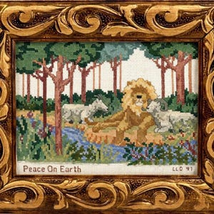 May include: Cross-stitch artwork in a gold ornate frame. The image depicts a lion and lamb resting peacefully in a forest setting, with the text "Peace On Earth" at the bottom. The artwork is detailed with various colors.