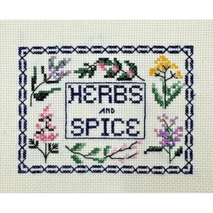 May include: Cross-stitch embroidery featuring the words "HERBS AND SPICE" in a blue rectangle, surrounded by a blue border. The design includes embroidered flowers in pink, yellow, and purple.