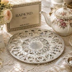 Puede incluir: Una bandeja redonda de metal plateado con un intrincado borde de celosía calada y un patrón central de rayos de sol. La bandeja se muestra junto a una tetera de porcelana floral, una taza a juego, una caja con la etiqueta RAIMOND 254 y un colador de té.