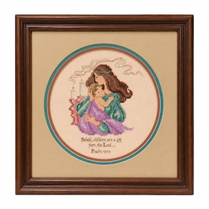 Puede incluir: Un bordado en punto de cruz enmarcado que muestra a una persona con cabello largo castaño sosteniendo a un bebé. La escena incluye dos velas encendidas y el texto "Behold, children are a gift from the Lord... Psalm 127:3". Obra en marco de madera oscura.