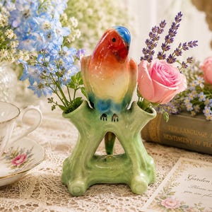 May include: A ceramic vase shaped like a green tree branch with a perched parrot featuring a red, blue, and white head and body. The vase has two openings for flowers, currently holding blue delphiniums and a single pink rose blossom.