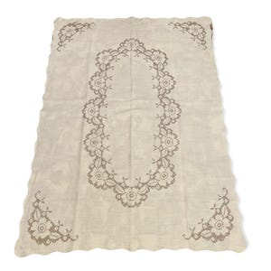 Puede incluir: Mantel rectangular color crema con un diseño bordado floral detallado. El bordado es de un tono crema más oscuro y presenta un gran patrón ovalado en el centro, con diseños florales más pequeños en cada esquina.