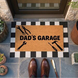 May include: A rectangular doormat with the text "DAD'S GARAGE" and tool silhouettes. The mat is bordered with a black and white checkered pattern. Brown leather shoes and a shoe horn are in the foreground.