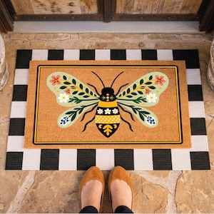 May include: A rectangular doormat with a large bee design. The bee has floral wings in shades of green, pink, and blue. The mat is on a black and white striped rug. The background includes a stone floor and a wooden door.