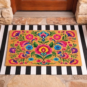 Puede incluir: Felpudo rectangular con un vibrante dise&ntilde;o floral en tonos rosa, azul, amarillo y morado. La alfombra est&aacute; sobre una alfombra a rayas blancas y negras, con plantas en macetas a ambos lados.