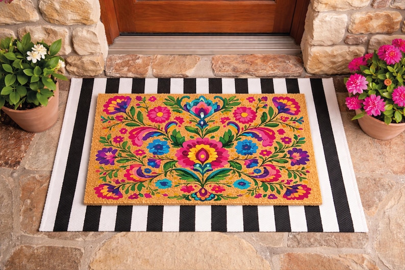 Puede incluir: Felpudo rectangular con un vibrante dise&ntilde;o floral en tonos rosa, azul, amarillo y morado. La alfombra est&aacute; sobre una alfombra a rayas blancas y negras, con plantas en macetas a ambos lados.