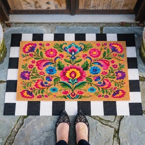 Puede incluir: Una alfombra rectangular con un vibrante dise&ntilde;o floral en tonos rosa, azul, amarillo y verde sobre un fondo beige. Se coloca sobre una alfombra a rayas blancas y negras. La escena est&aacute; ambientada al aire libre.