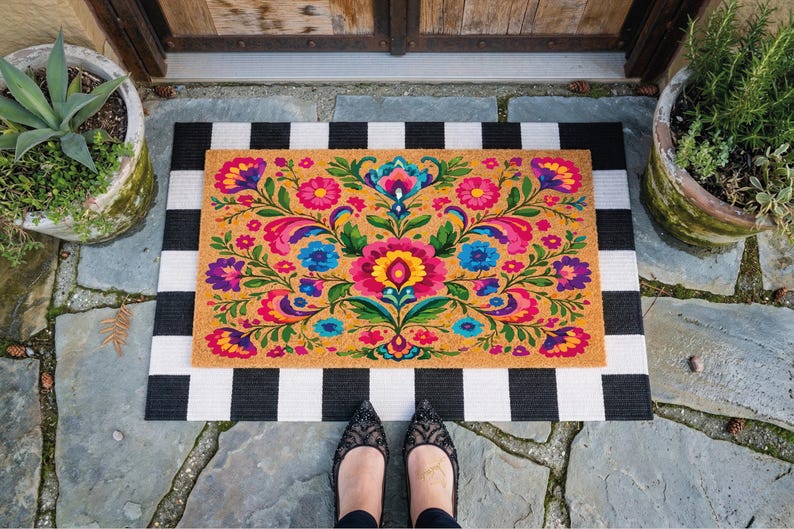 Puede incluir: Una alfombra rectangular con un vibrante dise&ntilde;o floral en tonos rosa, azul, amarillo y verde sobre un fondo beige. Se coloca sobre una alfombra a rayas blancas y negras. La escena est&aacute; ambientada al aire libre.