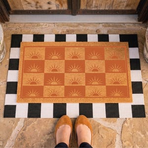 May include: A rectangular doormat with a checkerboard pattern in orange and white, featuring a repeating sun design. The mat is layered over a black and white striped rug. The scene is set on a stone patio, with potted plants on either side.