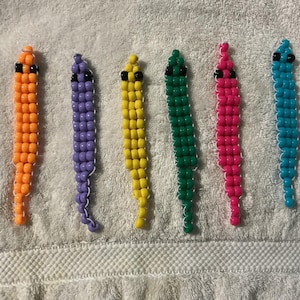 May include: Six colorful beaded worm-like figures in orange, purple, yellow, green, pink, and blue. Each has black bead eyes and a white beaded outline. The figures are lying on a white, textured surface.