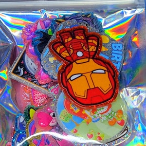 May include: A collection of assorted colorful stickers with glittery finishes, including a prominent red and gold Iron Man character design, all contained within a clear, iridescent, holographic resealable plastic storage pouch.