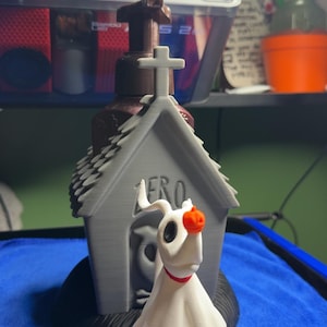 May include: A 3D printed grey doghouse with a cross on top and the word ZERO embossed on the front. A white ghost dog figure with a red collar and a small orange pumpkin nose is positioned in front of the doghouse, which sits on a black base.