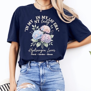 Puede incluir: Camiseta azul marino con un diseño floral de hortensias azules, rosas y moradas. El texto dice "IN MY NT lady LAY ERA" y "Hydrangea Lover Floral Garden Bloom".