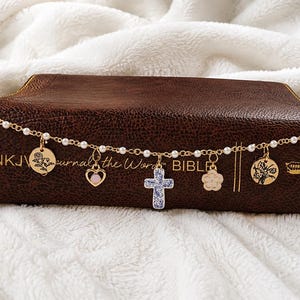 May include: A gold-toned chain bracelet featuring small white faux pearls and multiple charms, including a blue and white patterned cross, a heart with a pink stone, a flower, and two round discs with floral engravings, plus two rectangular tags.