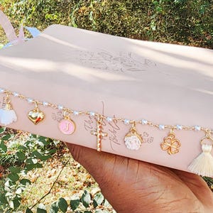 May include: A light pink Bible featuring a decorative gold-tone chain draped across the cover. The chain is adorned with several charms, including a white tassel, a gold heart, a pink circular charm with the letter J, a cross, a white flower, a four-leaf clover, and a cream tassel.