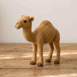 Op de afbeelding: Een met naaldvilt gemaakte kameelfiguur van beige wol. De kameel heeft één bult, een lange nek, een textuurrijke vacht, donkere kraaloogjes en donkerbruine wollen accenten op de onderbenen en voeten, staand in een profielpositie.
