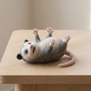 May include: A needle-felted opossum figurine lying on its back with its paws raised. The creature features a white belly, grey fur on its back and head, black ears, a pink nose, and a long, smooth, light pink tail extending from its body.