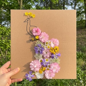 May include: A floral peacock design on a square piece of cardboard. The peacock is outlined in black, with a tail made of pink, yellow, white, and purple flowers. The artwork is held by a hand against a green backdrop.