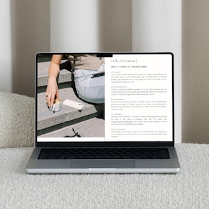 May include: A silver laptop displaying a webpage with text and an image of a person holding a coffee cup and a phone. The laptop is on a white surface, with a textured pillow and a white wall in the background.