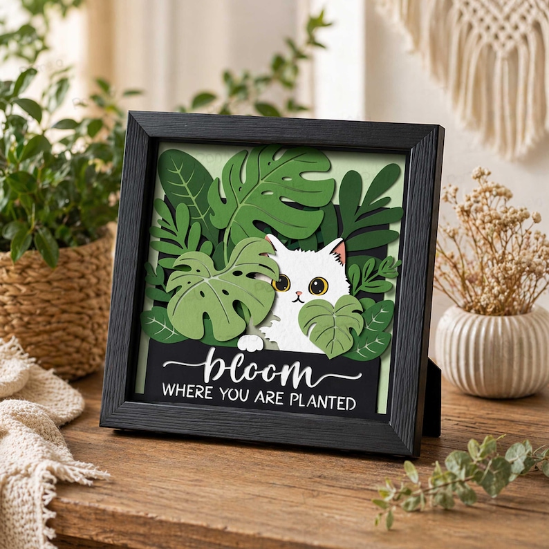 May include: A framed artwork featuring a white cat peeking through green leaves. The black frame holds a layered design with a light green background. The text "bloom where you are planted" is displayed on a black banner.