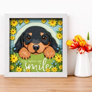 May include: A framed 3D paper art piece featuring a dachshund dog surrounded by yellow flowers. The artwork has a white frame and the text "YOU MAKE ME smile" in cursive. A vase of tulips sits to the right.