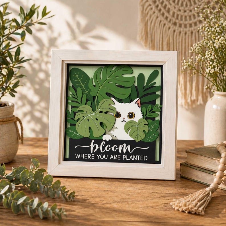 May include: A framed artwork featuring a white cat peeking through green tropical leaves. The artwork includes the text "bloom where you are planted" on a black background, set within a white frame. The artwork is on a wooden surface.