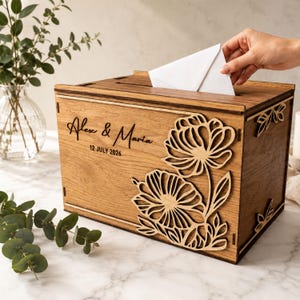May include: A rectangular wooden card box with a slot on top, featuring a laser-cut floral design on the front panel. The wood is engraved with the text Alex & Maria and the date 12 JULY 2026. The box has a natural brown finish.