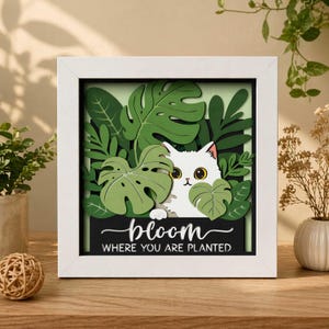May include: A framed, layered paper art piece featuring a white cat peeking through green leaves. The artwork includes the words "bloom where you are planted" on a black background, all within a white frame. The cat has yellow eyes.