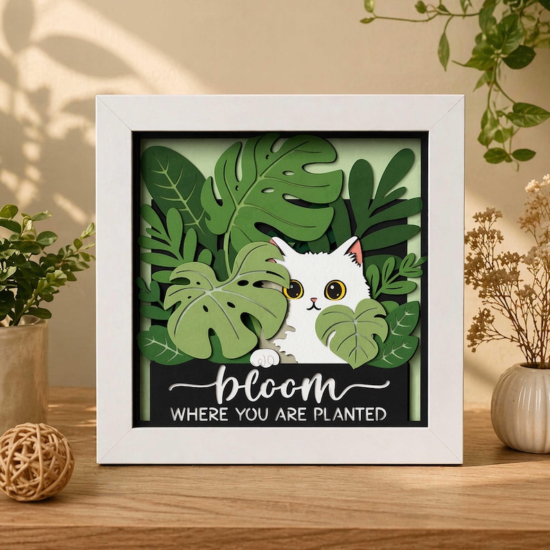 May include: A framed, layered paper art piece featuring a white cat peeking through green leaves. The artwork includes the words "bloom where you are planted" on a black background, all within a white frame. The cat has yellow eyes.