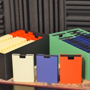 May include: A black storage box filled with cream-colored dividers and a green storage box filled with blue dividers. In front are three individual rectangular card cases in cream, purple, and orange, each featuring a top cutout for easy access.