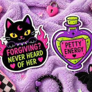 May include: A collection of stickers and accessories with a cute, gothic theme. Includes a black cat sticker with the text "Forgiving? Never Heard of Her", a purple potion sticker with "Petty Energy", and a plush bat.
