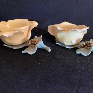 May include: Two ceramic rose-shaped bowls with a light yellow glaze. Each bowl is sitting on a leaf-shaped base with a small brown teddy bear figurine.