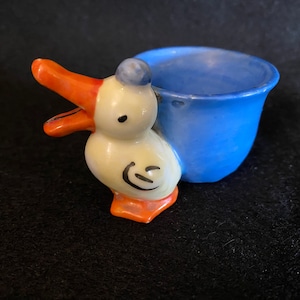 May include: A blue and white ceramic egg cup shaped like a duck. The duck has an orange beak and feet.