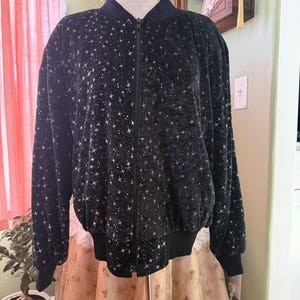 May include: A black bomber jacket featuring a repeating pattern of small white stars and sparkles. The jacket has a solid black ribbed collar, cuffs, and hem, with a full-length front zipper closure. The fabric appears to have a soft, velvet-like texture.