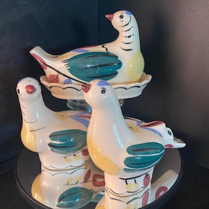 May include: Three ceramic bird figurines with colorful hand-painted details. The birds are white with teal, yellow, red, and blue accents. One bird sits on a decorative plate. The figurines are displayed on a mirrored surface.