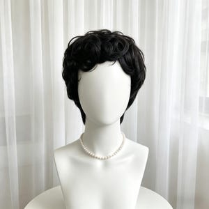 Könnte beinhalten: Eine schwarze Perücke mit kurzem, lockigem Haar wird auf einem weißen Schaufensterkopf präsentiert. Die Schaufensterpuppe trägt eine zarte Perlenkette. Der Hintergrund zeigt einen weißen Vorhang, der ein weiches, diffuses Licht erzeugt.