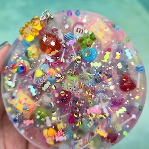 May include: A circular clear resin pendant filled with colorful miniature candy charms, including lollipops, gummy bears, and chocolate pieces, accented with iridescent glitter and small beads throughout the translucent material.