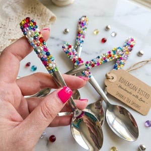 May include: A set of silver-toned metal tea spoons with handles embellished with multicolored rhinestones. A brown paper tag is attached, featuring the printed text: BOHEMIAN BISTRO - Handmade with Love - SPARKLE TEA SPOONS. The spoons have a polished finish.