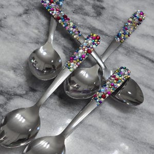 May include: A set of four silver-toned metal spoons featuring handles embellished with a dense arrangement of multicolored, iridescent, and clear rhinestones in various sizes, creating a vibrant, textured, and reflective surface on each spoon.