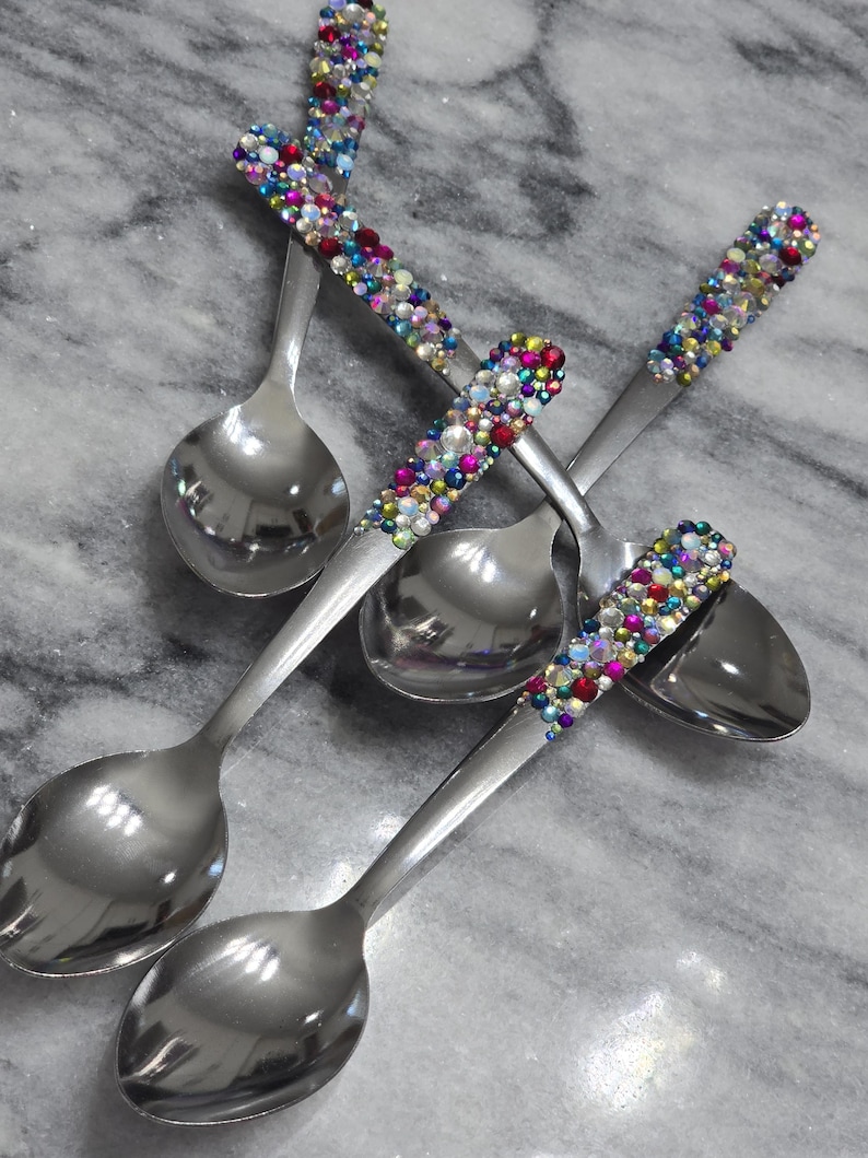 May include: A set of four silver-toned metal spoons featuring handles embellished with a dense arrangement of multicolored, iridescent, and clear rhinestones in various sizes, creating a vibrant, textured, and reflective surface on each spoon.