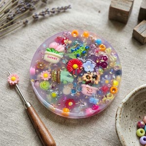 May include: A circular, translucent purple resin coaster embedded with colorful miniature charms, including flowers, potted plants with the text Sweet and Smile, and various beads. A metal tool with a wooden handle and a small pink flower topper is adjacent.