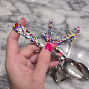May include: A set of silver-toned metal spoons featuring handles densely encrusted with a variety of colorful, multi-sized rhinestones in shades of pink, blue, yellow, green, and clear, creating a vibrant, textured, and reflective surface.