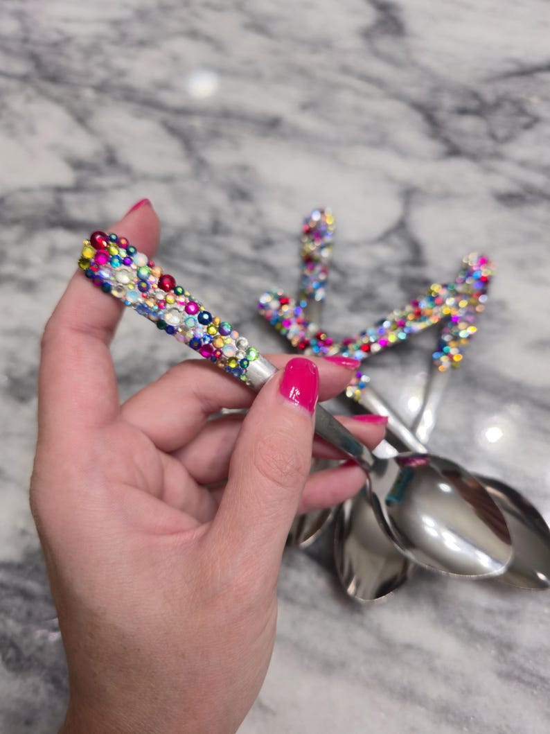 May include: A set of silver-toned metal spoons featuring handles densely encrusted with a variety of colorful, multi-sized rhinestones in shades of pink, blue, yellow, green, and clear, creating a vibrant, textured, and reflective surface.